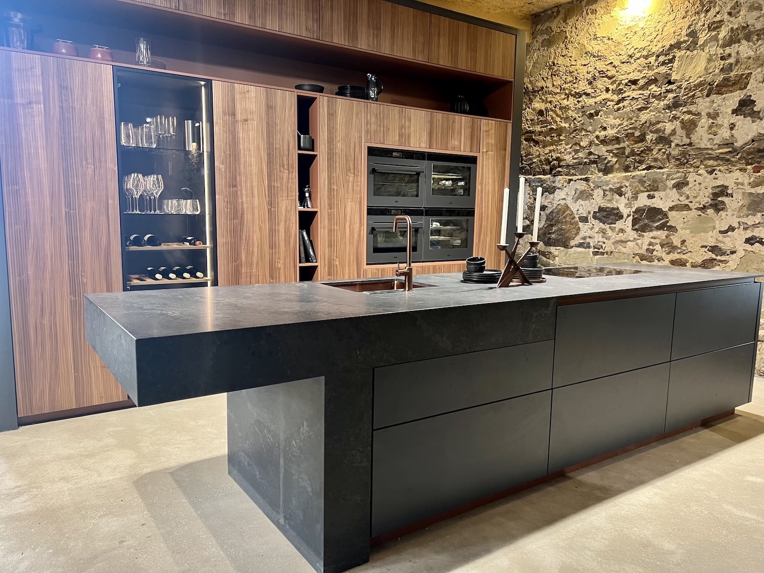 Moderne Küchenzeile mit grauer Oberfläche vor einer Steinwand und einem Holzimitat-Regal